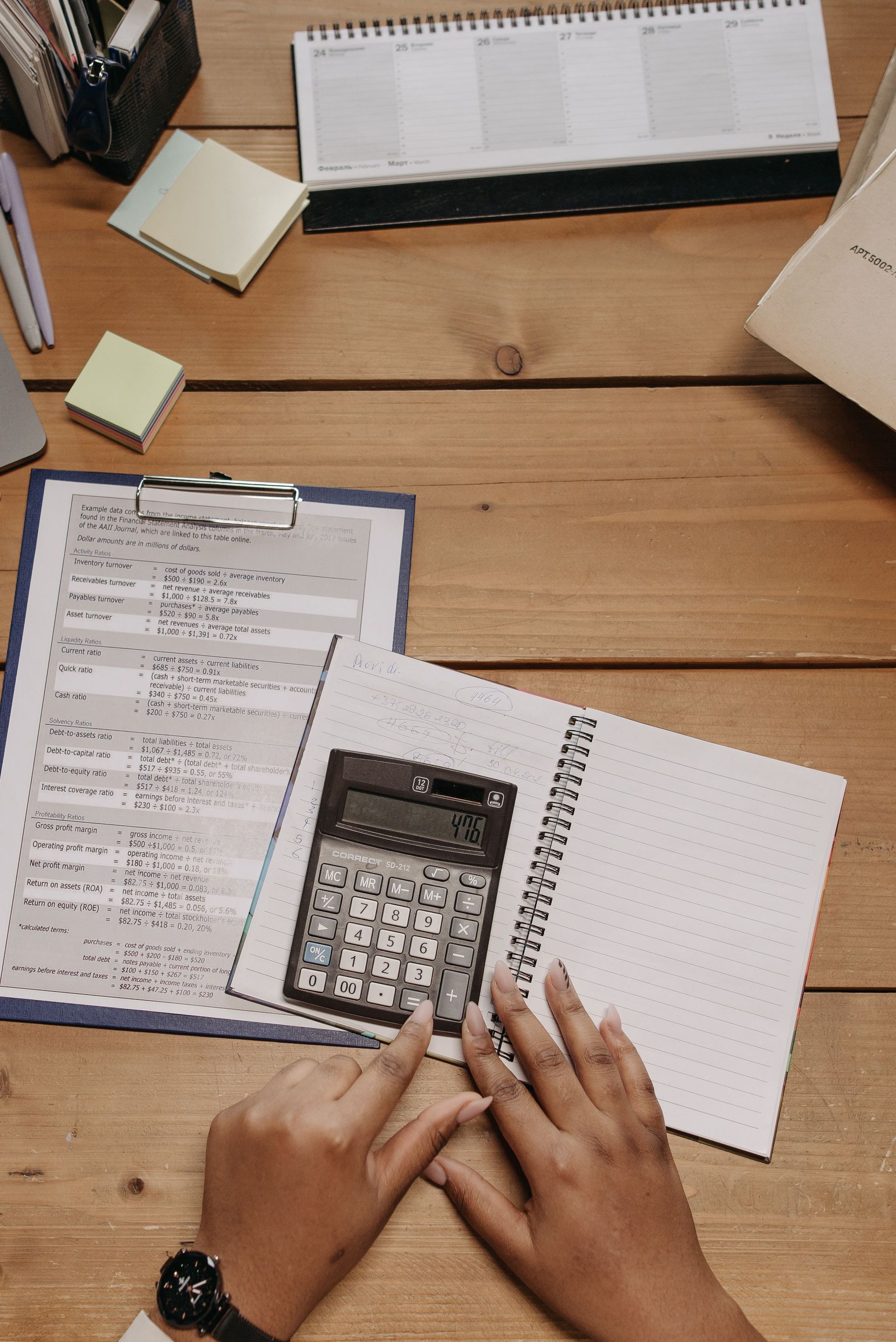 Top view of hands using calculator on office desk surrounded by documents and sticky notes.